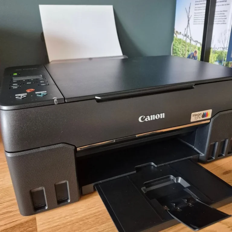 Canon MegaTank printer with paper loaded and its output tray open, on a wooden surface against a sage green wall.